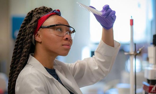 Biology student working in science lab in lab coat, with goggles, and looking at specimen